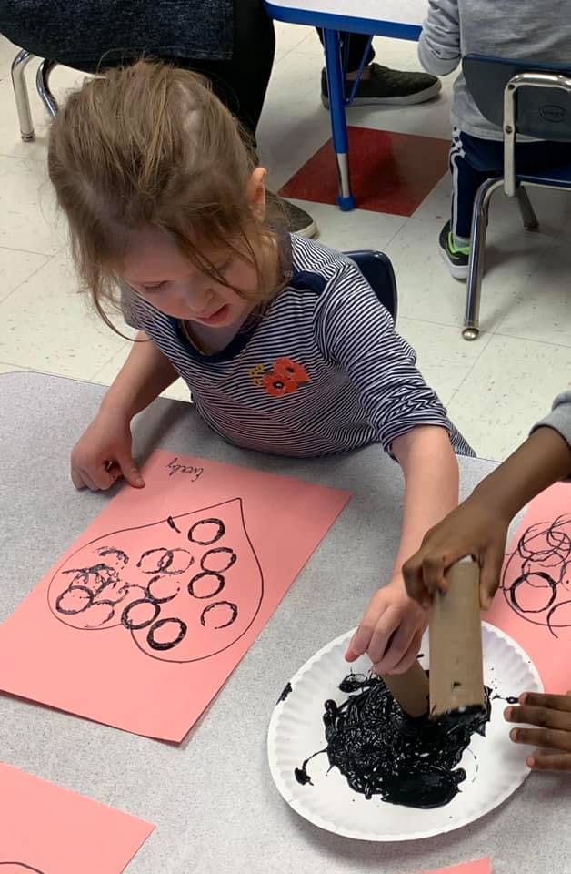 Child looking at a pink paper with black circles, another child dipping a tube in paint