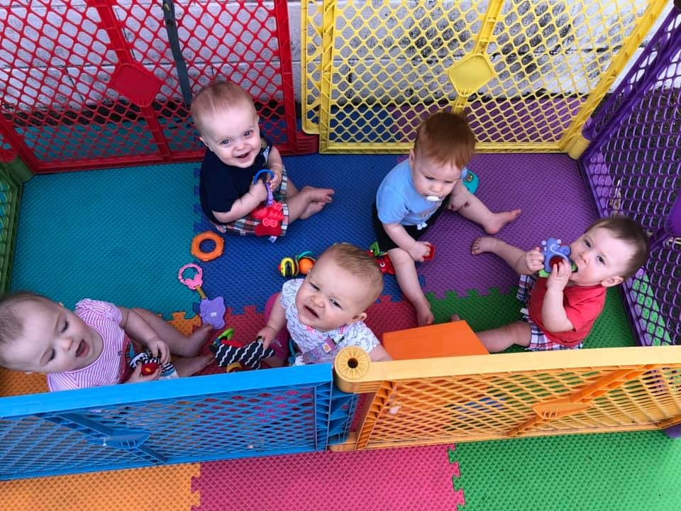 Five babies playing with toys in a colorful playpen