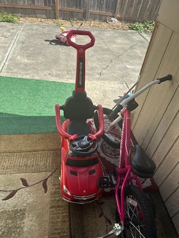 Red ride-on toy car with a push handle next to a pink bicycle