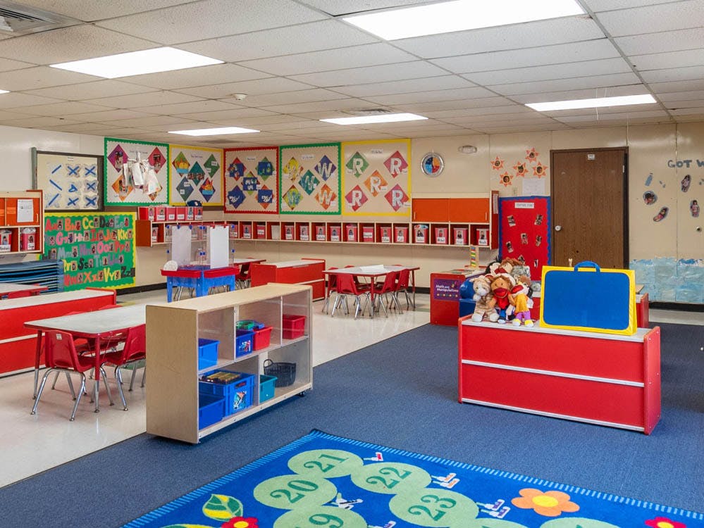 Empty classroom with small red tables and chairs, and a blue rug with numbers