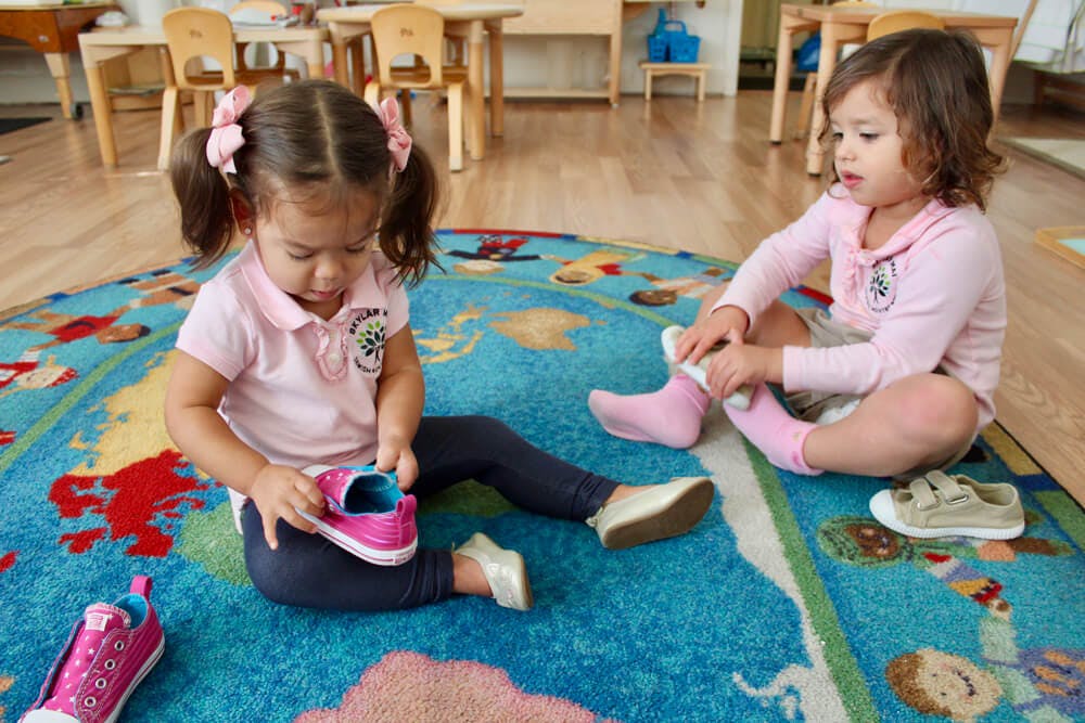 Two children sitting on a rug, one holding a shoe and the other putting on a sock