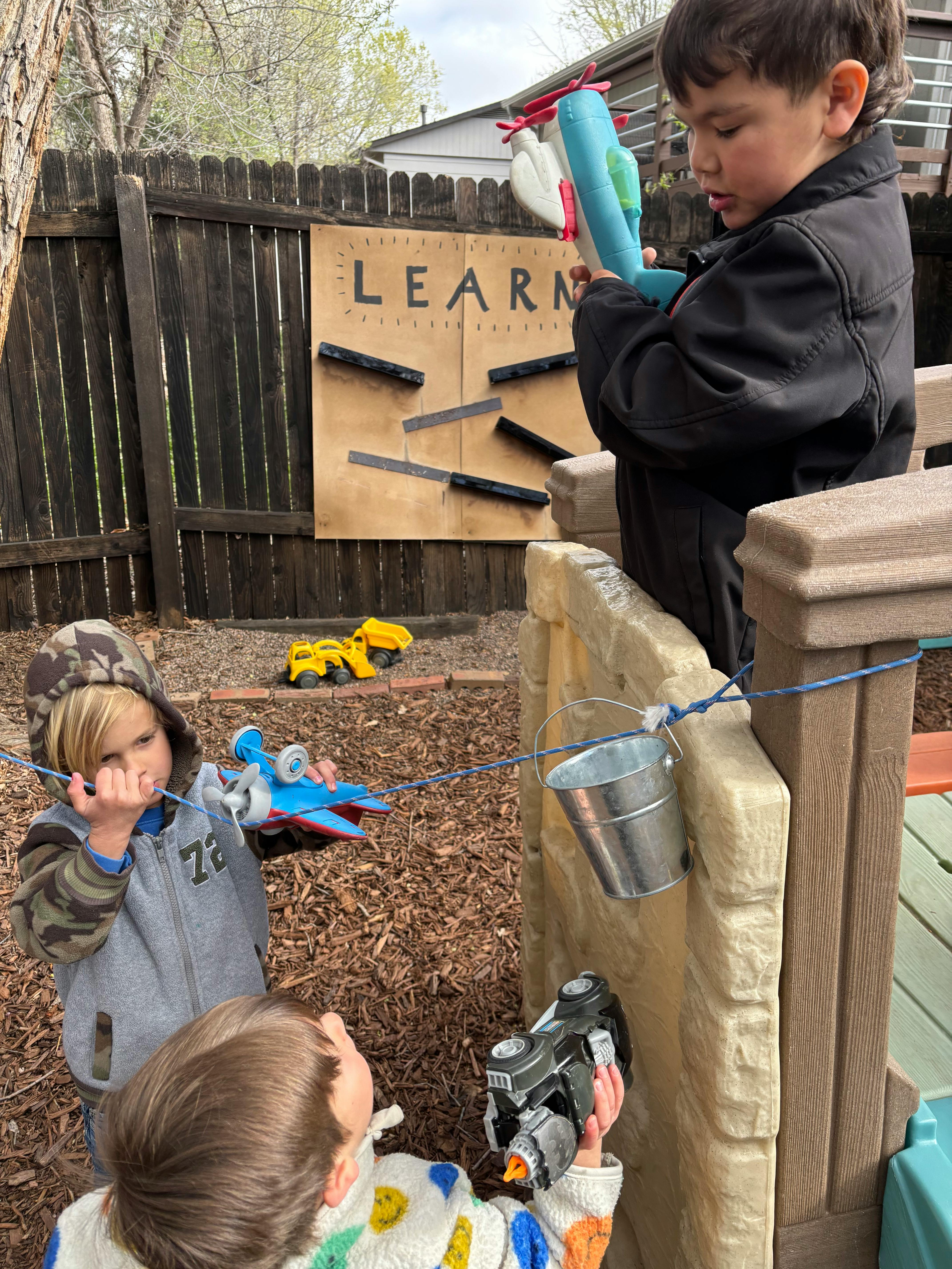 Three children playing with toys outdoors, one holding a toy plane