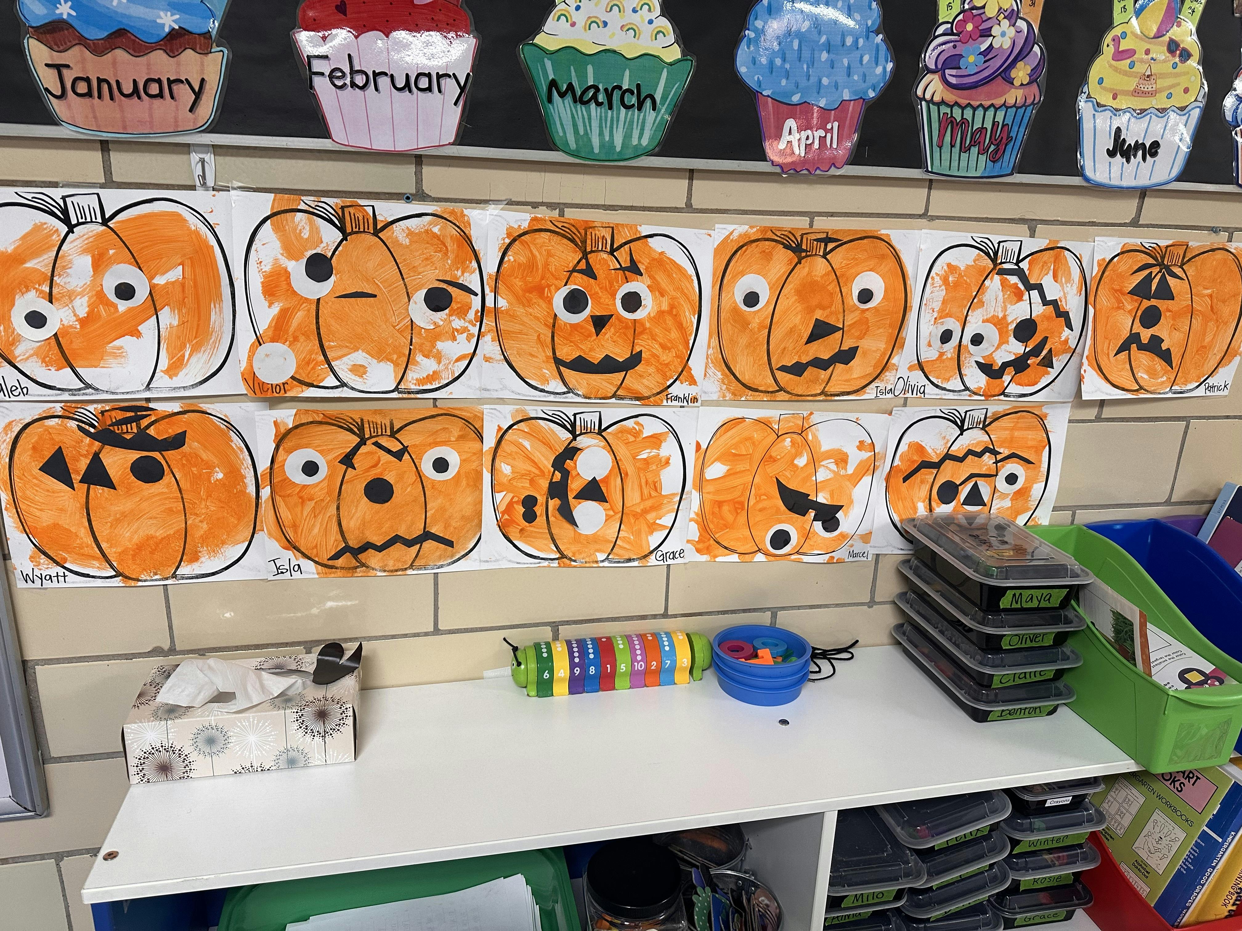 Children's pumpkin drawings displayed on a wall above a white shelf
