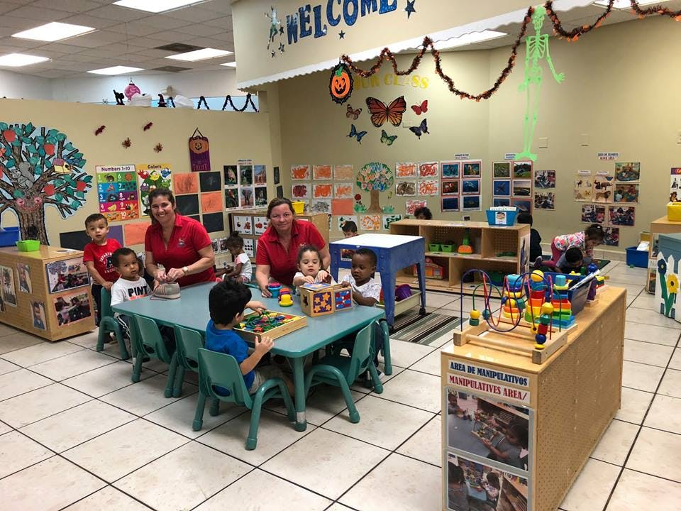 Two adults and several children playing with toys at tables in a classroom