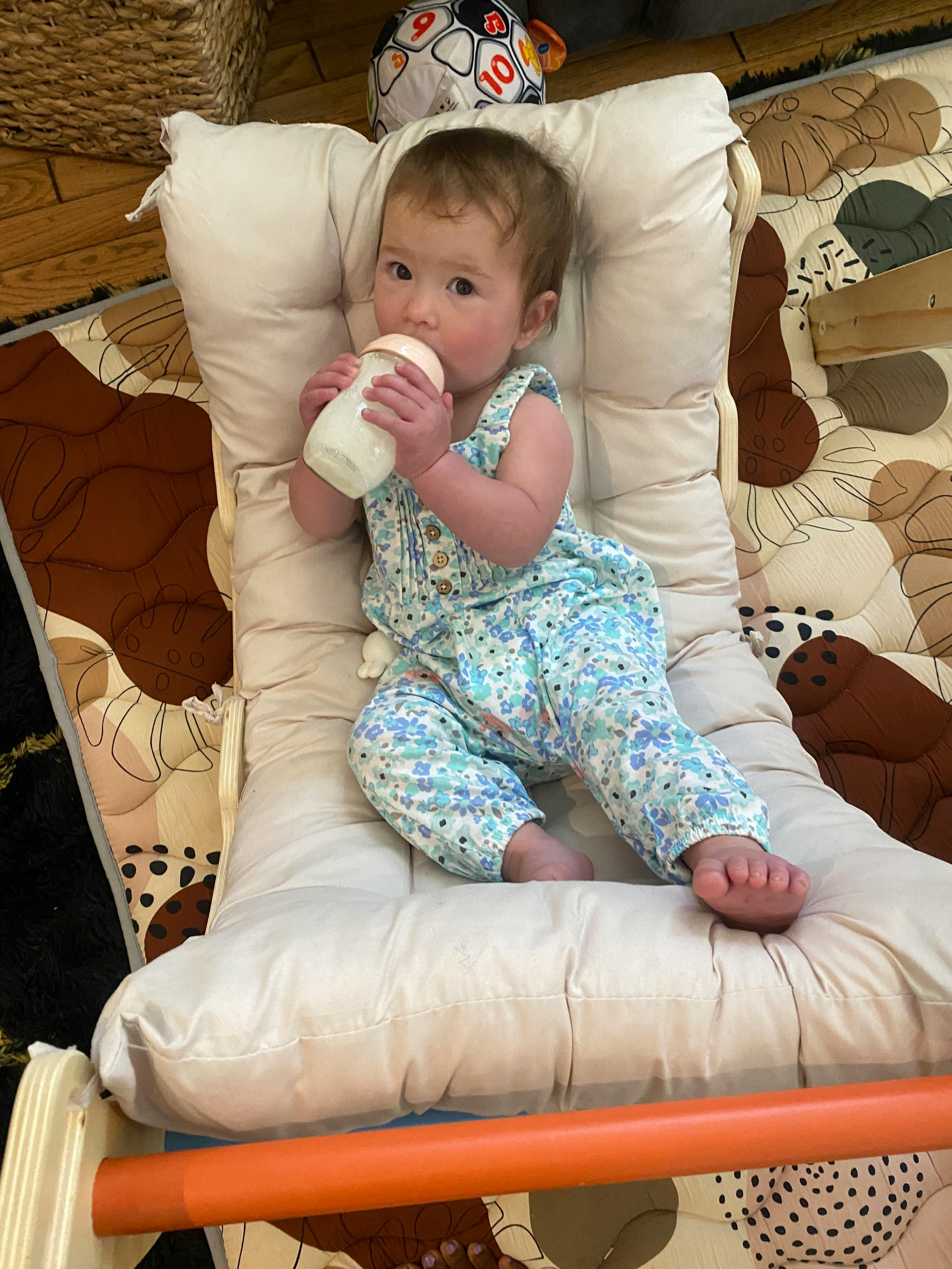 Child in a floral romper lying on a patterned mat and holding a bottle