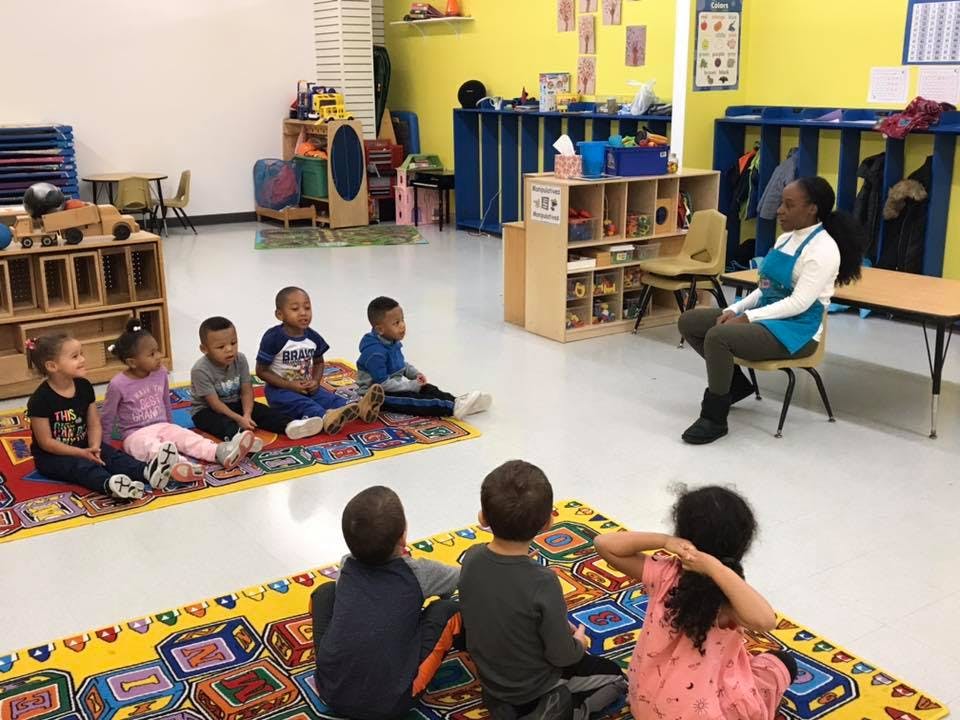 Adult sitting on a chair with children sitting on rugs in a classroom