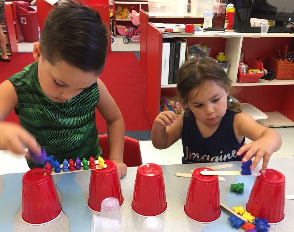 Two children playing with red cups and colorful small figures in a classroom