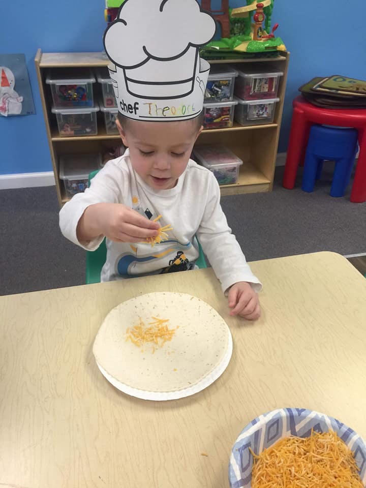 Child wearing a chef hat adding shredded cheese to a tortilla