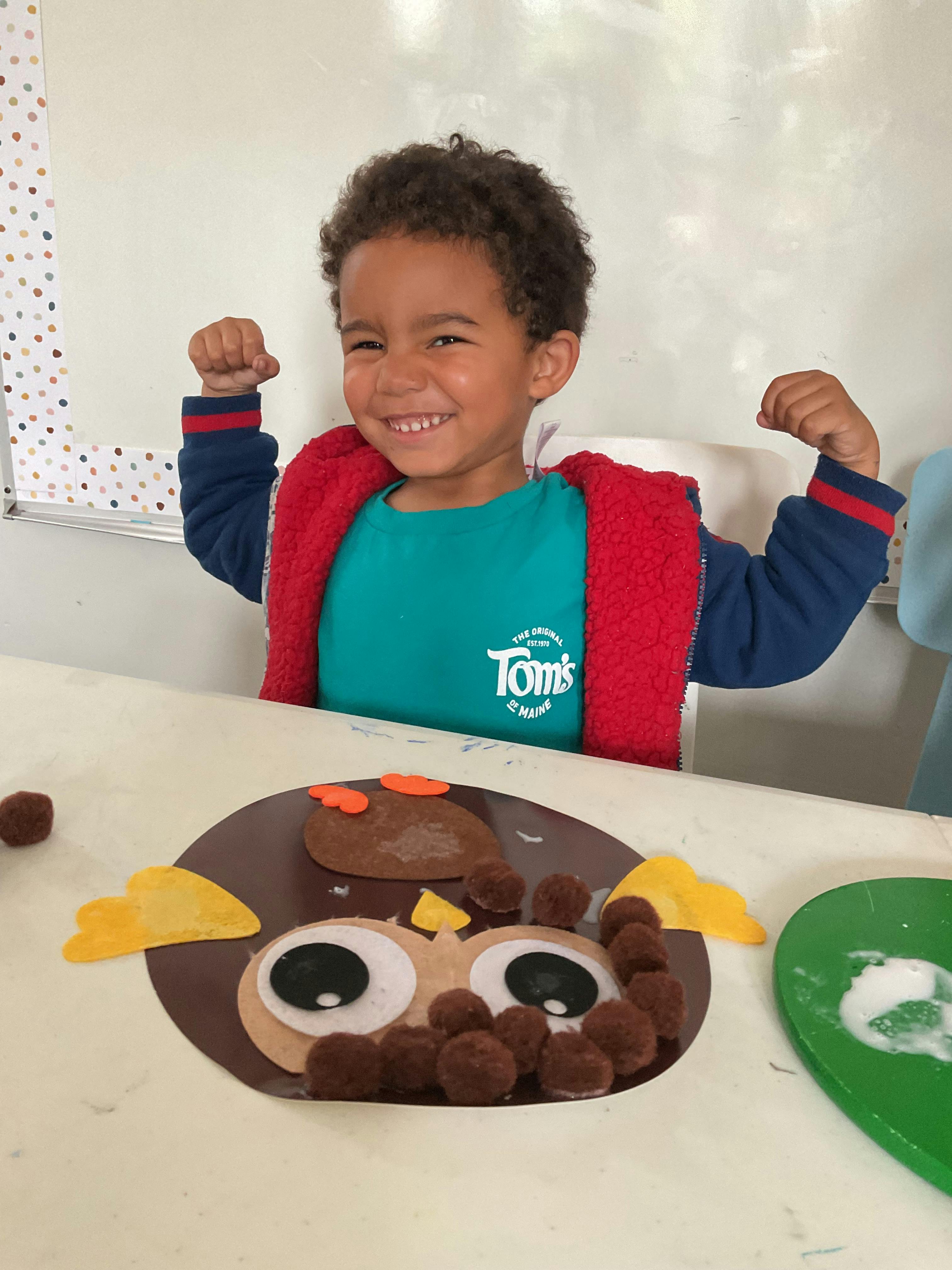 Child flexing muscles next to a handmade owl craft at a table