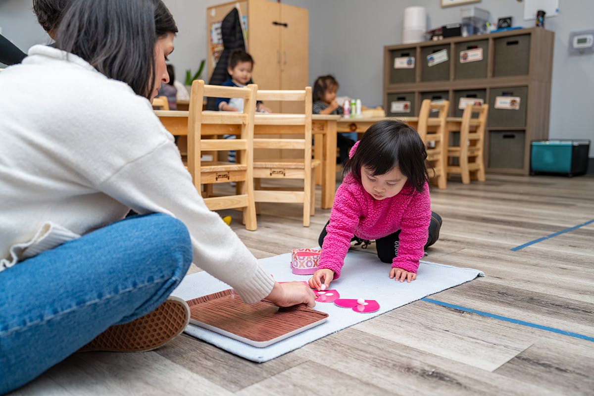 Person and child playing with pink shapes on the floor in a classroom