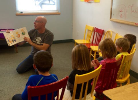 Person kneeling and reading a book to children in a classroom