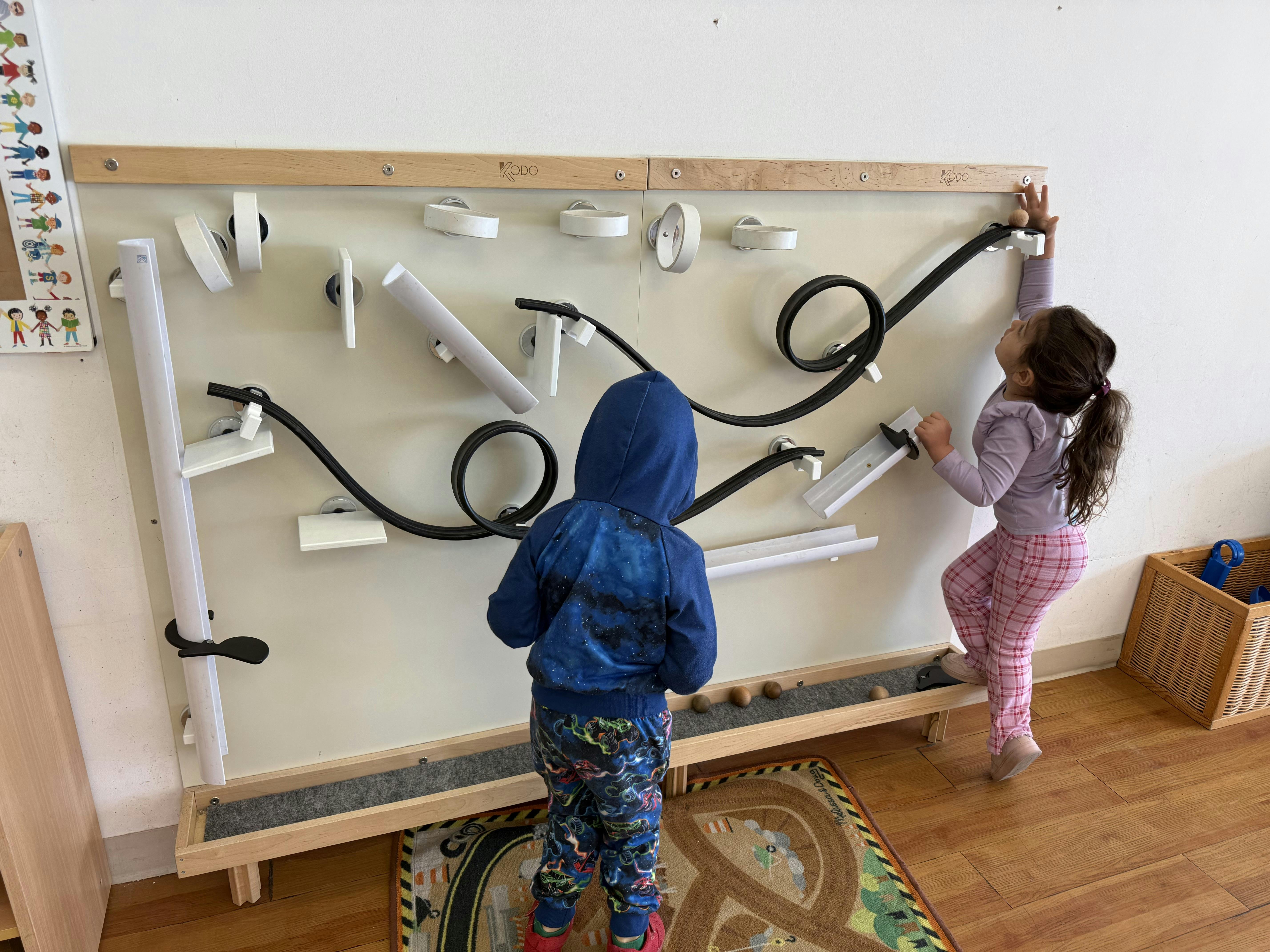 Two children playing with a marble run wall toy, with one child reaching up