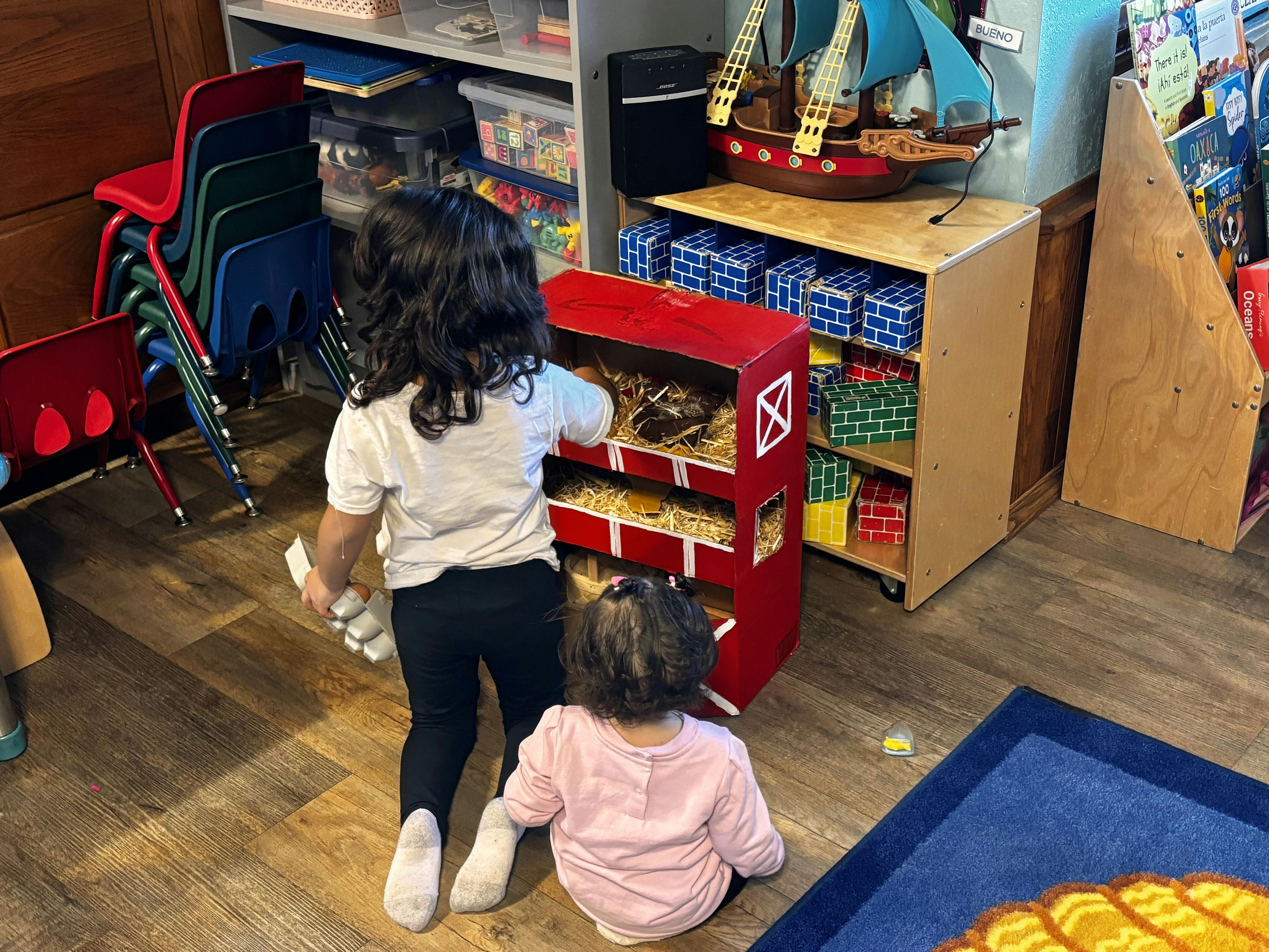 Two children playing with a red toy barn in a classroom