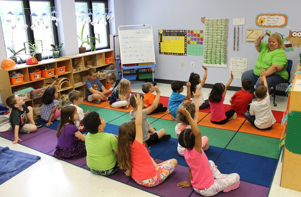 Adult teaching children in a classroom, with some children raising hands