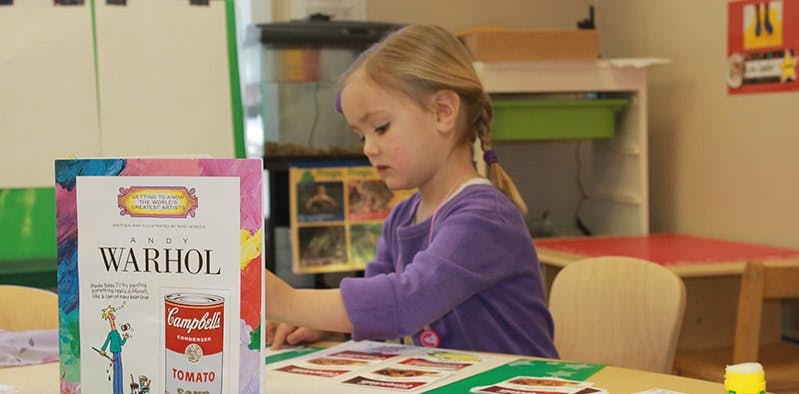 Child at a table with a book titled "WARHOL" and a Campbell's soup can