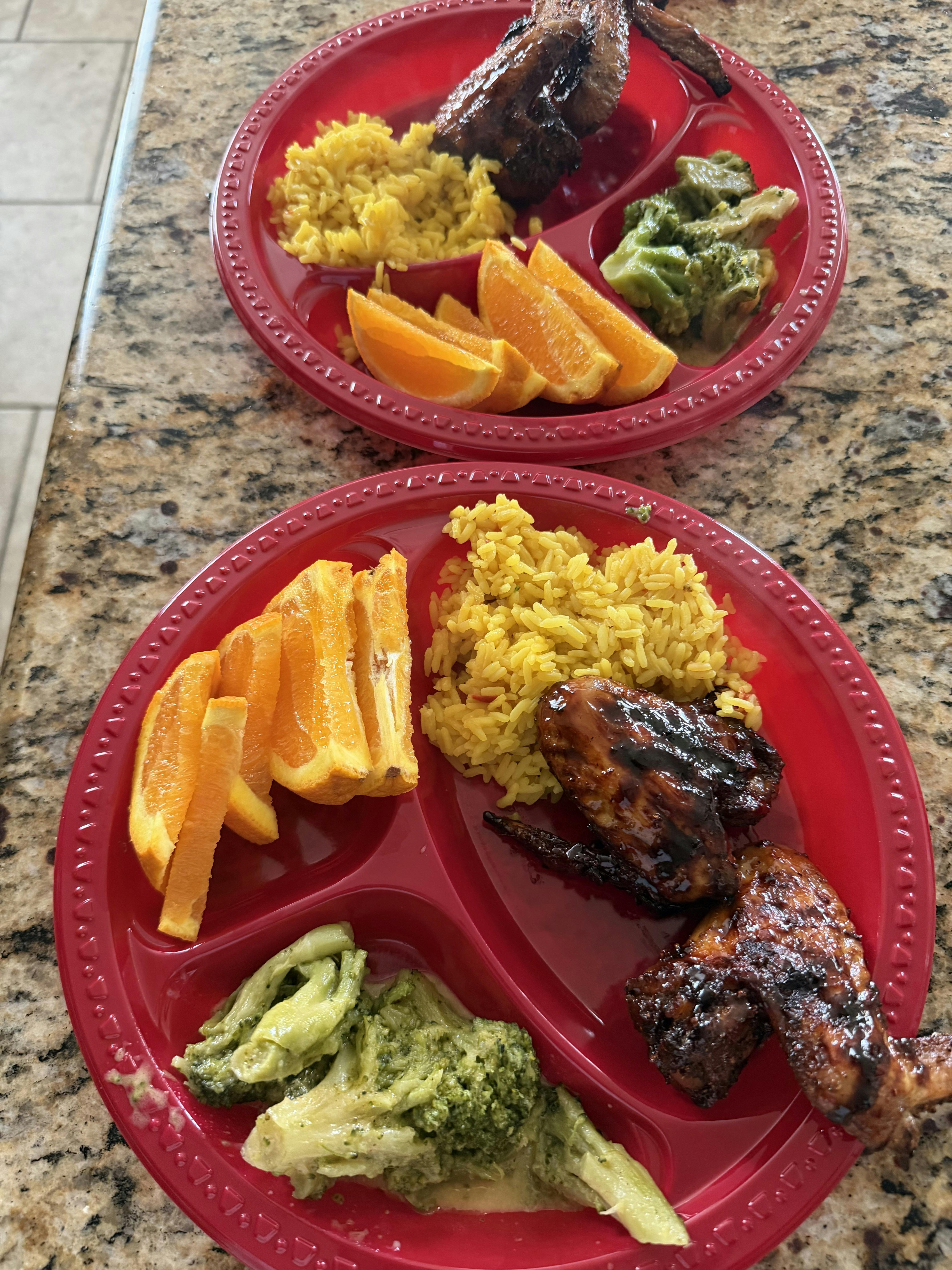 Two red partitioned plates containing glazed chicken wings, rice, and oranges
