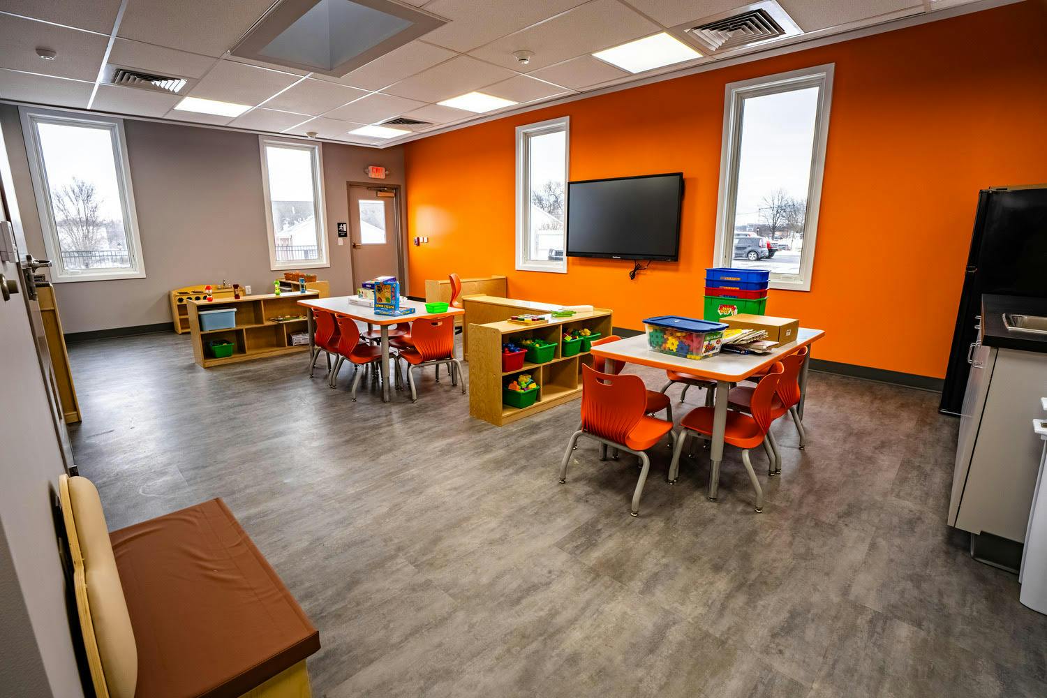 Classroom with small tables and chairs, shelves with toys, and orange accent walls