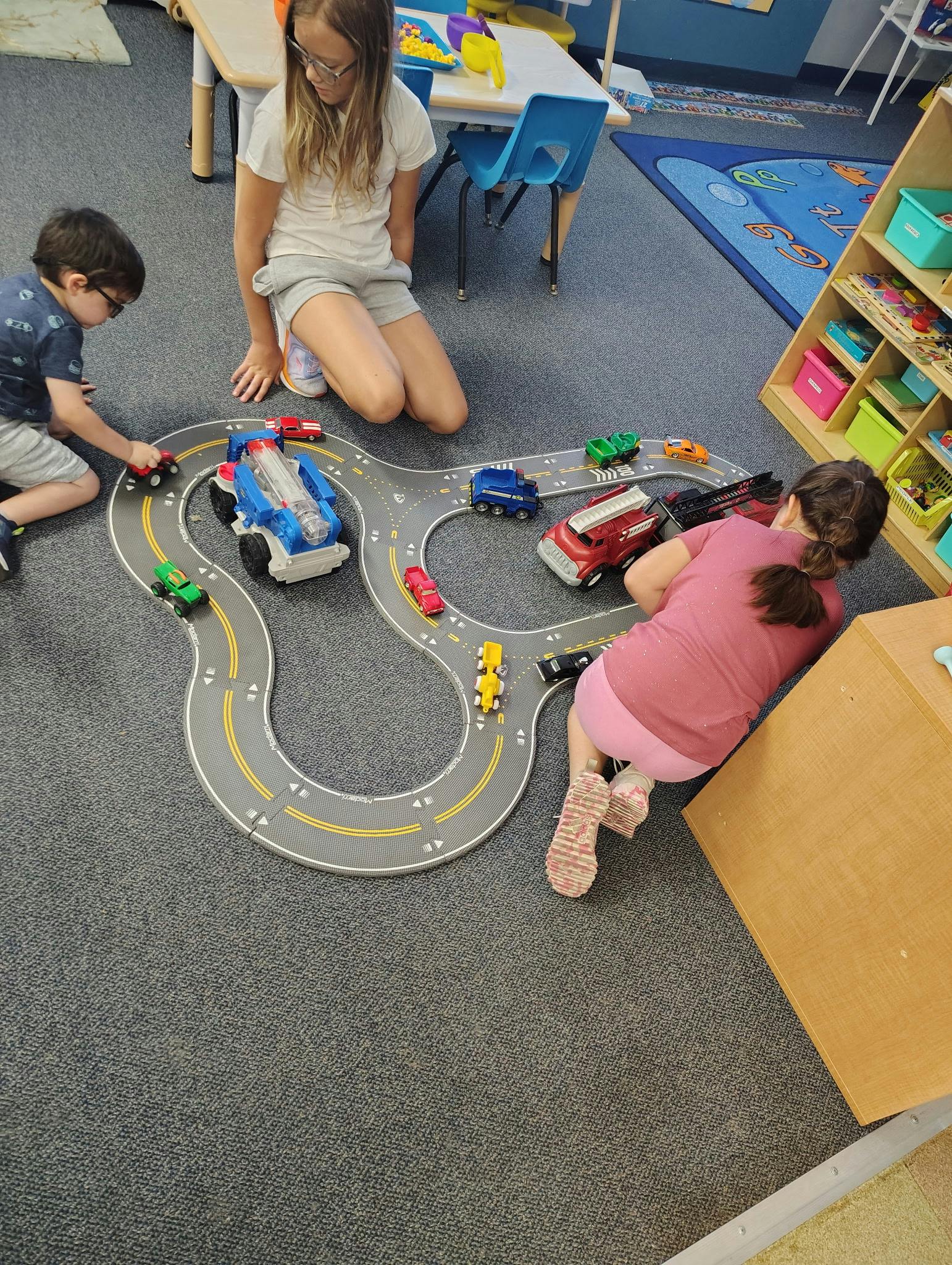 Three children playing with toy cars on a road mat in a classroom