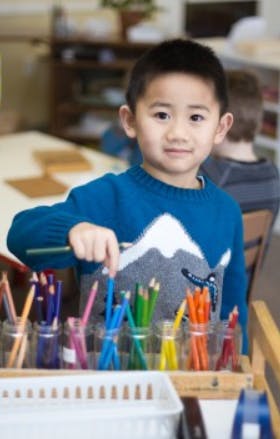 Child in a classroom selecting a colored pencil from a holder