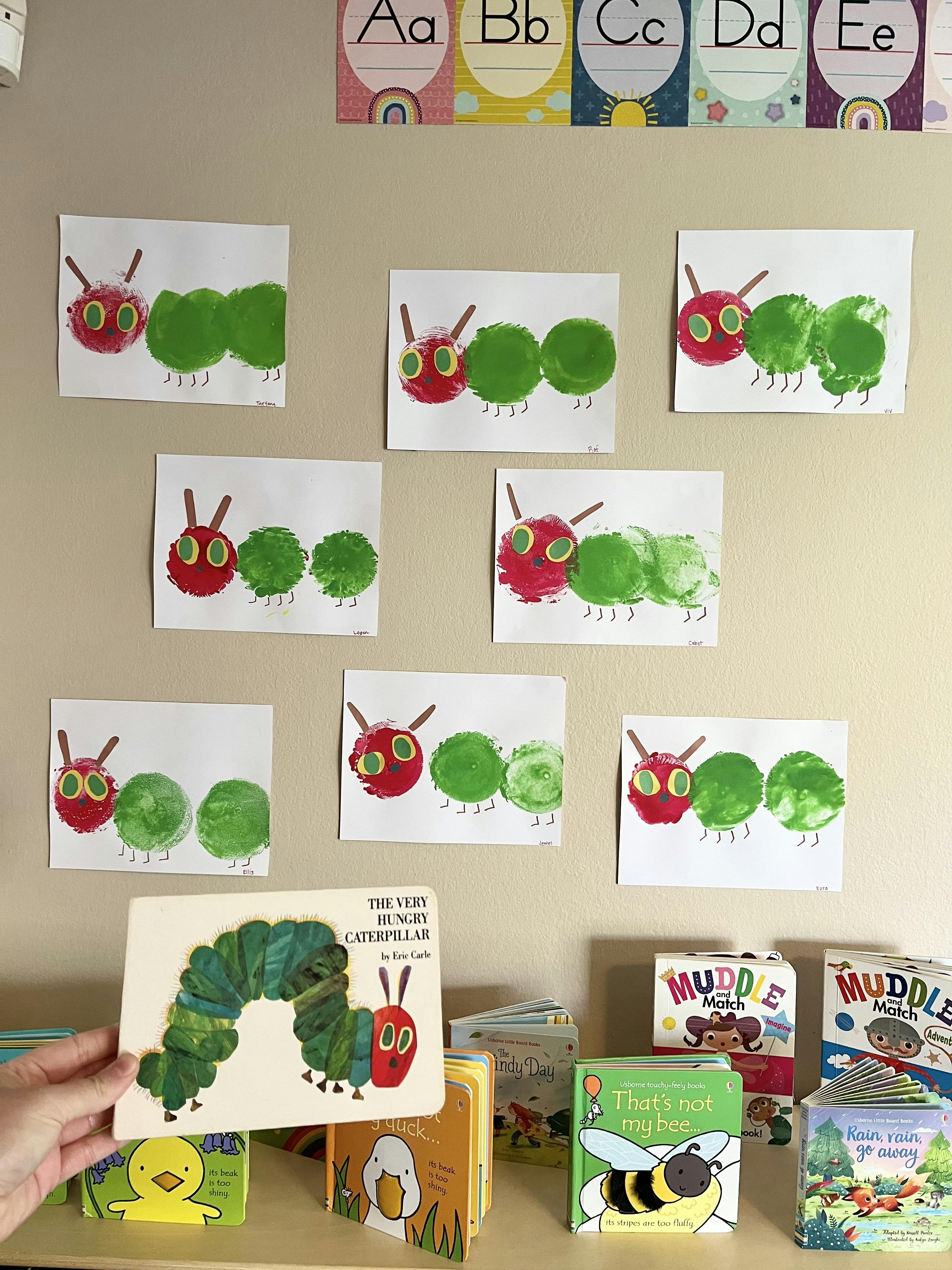 Person holding a book with a caterpillar on the cover, with caterpillar artwork on the wall