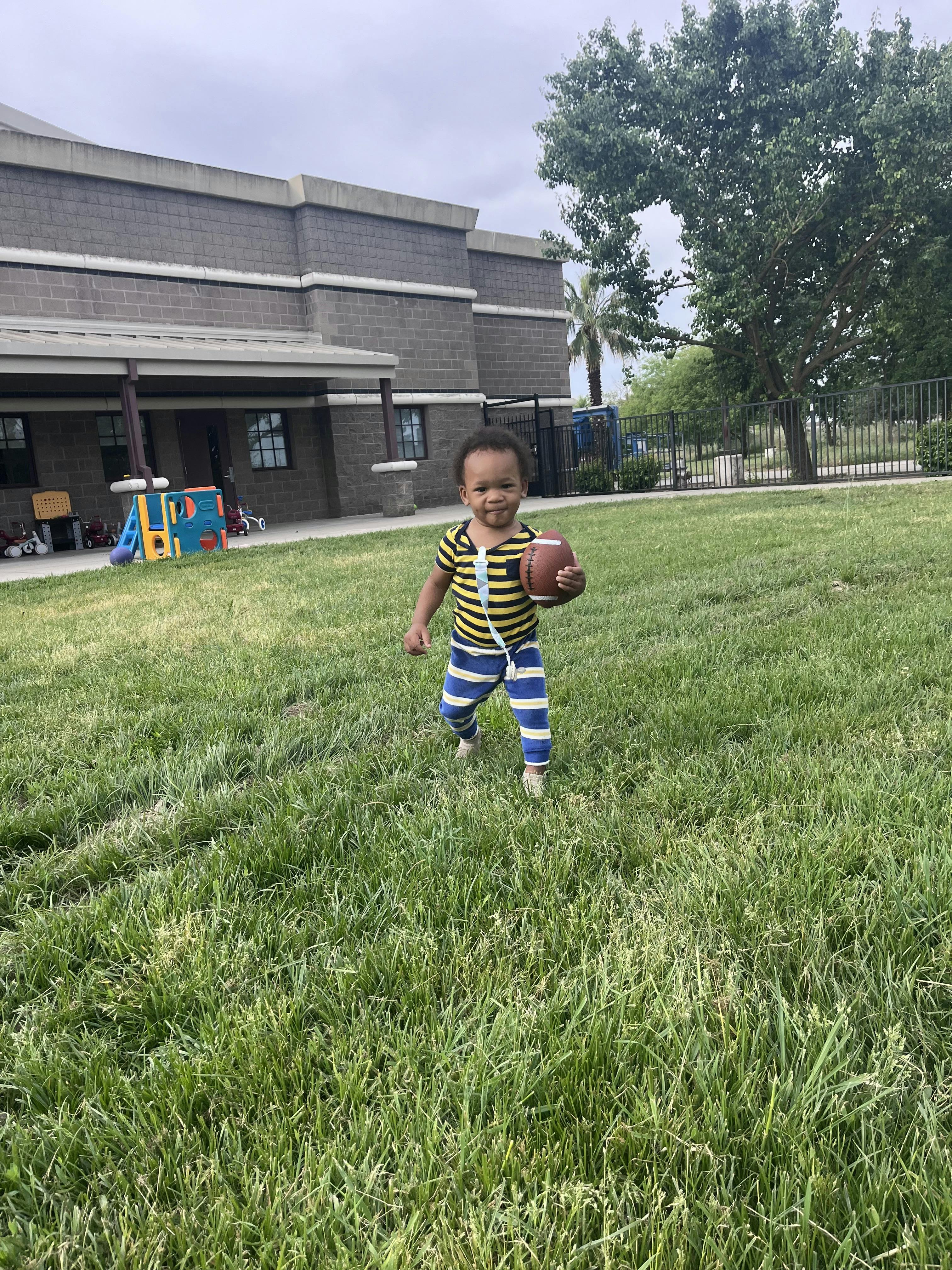 Child in a striped shirt and pants holding a football on a grassy field