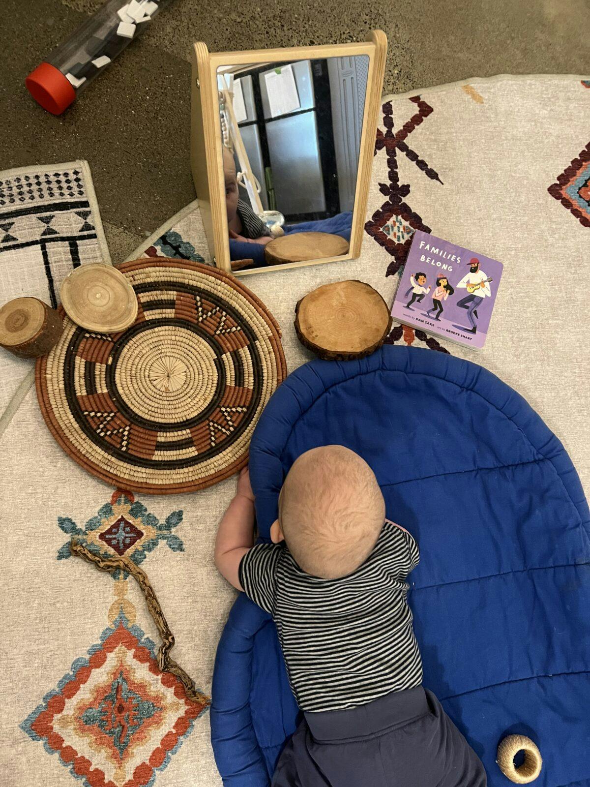 Baby lying on a blue mat, facing a mirror with a book titled "Families Belong" nearby