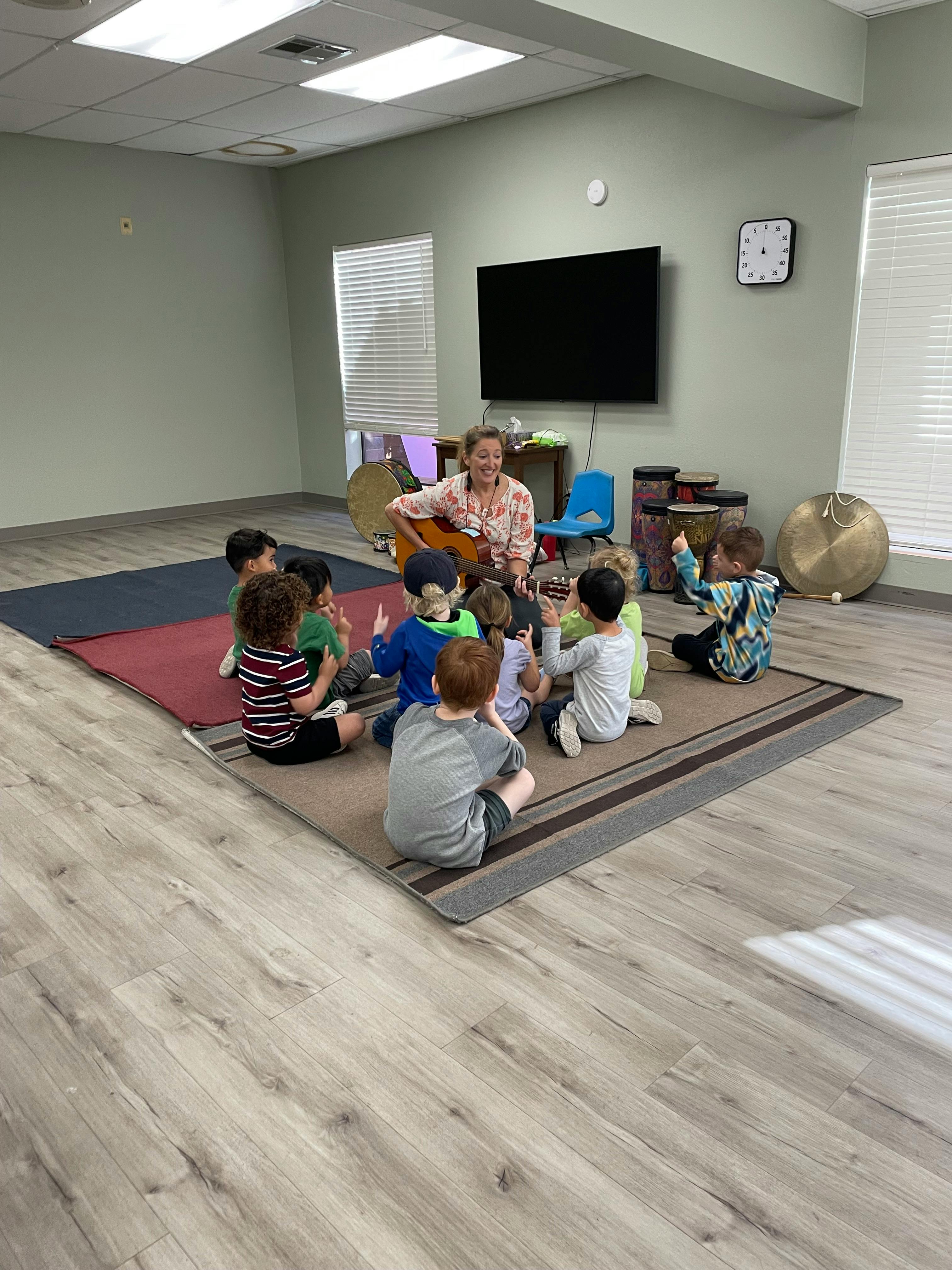 Person playing guitar for a group of children sitting on a rug