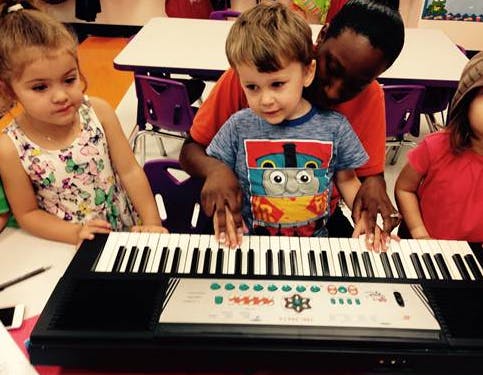 Adult helping a child play a keyboard, with another child watching