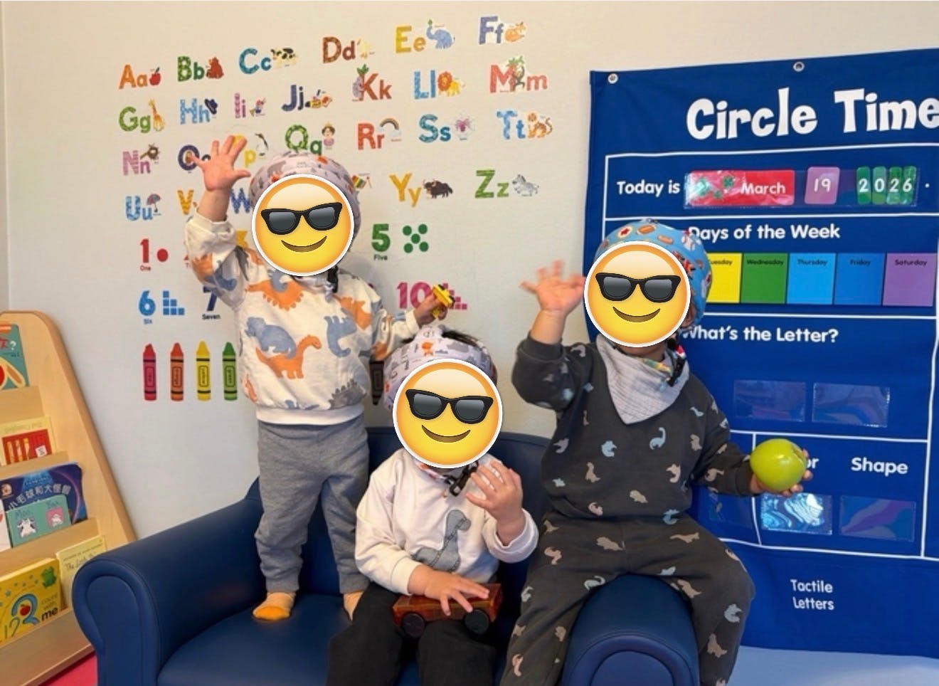 Three children in a classroom with alphabet letters on the wall, one holding an apple