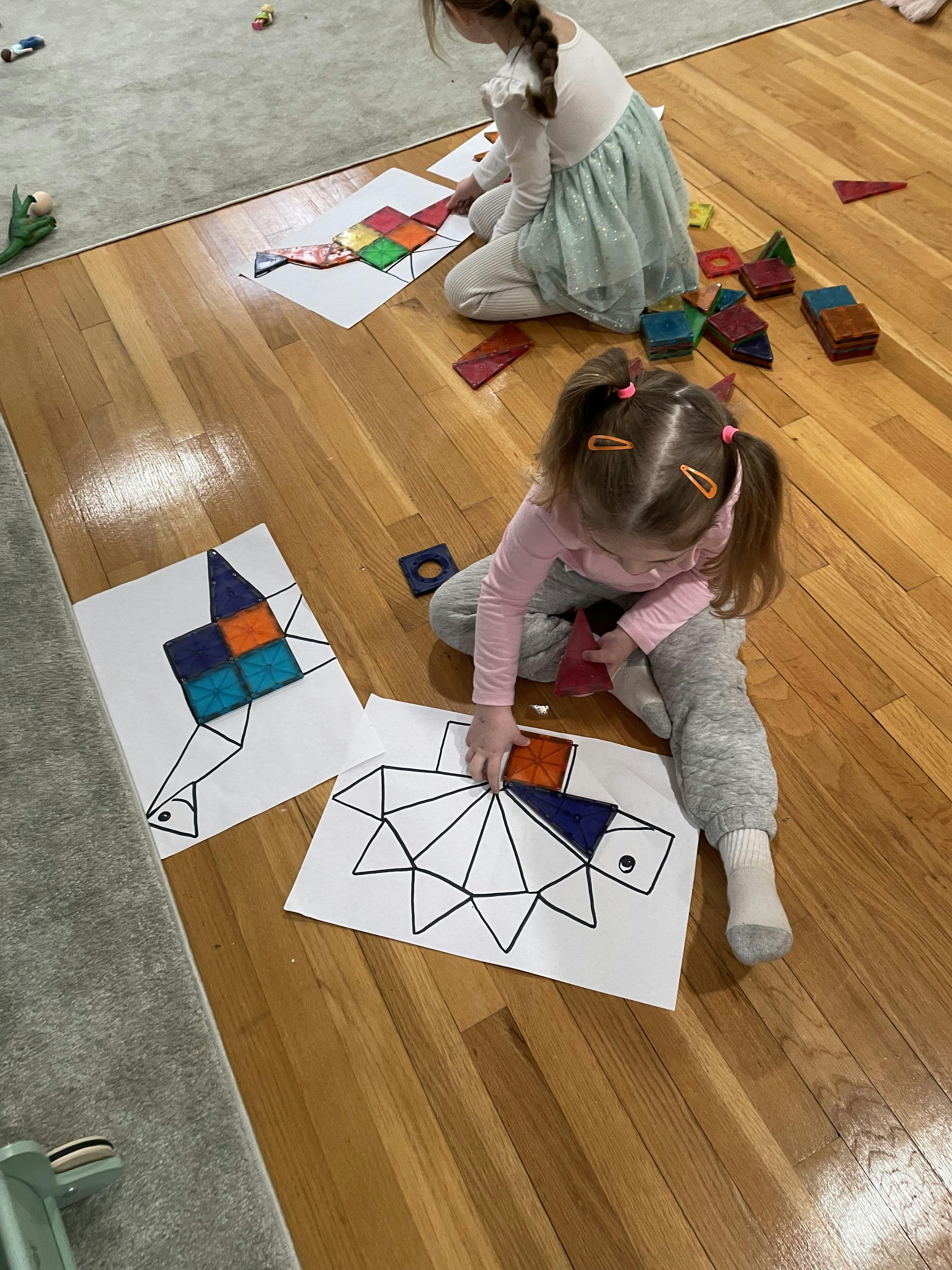Two children playing on a wooden floor, filling outlines on paper with colorful magnetic tiles