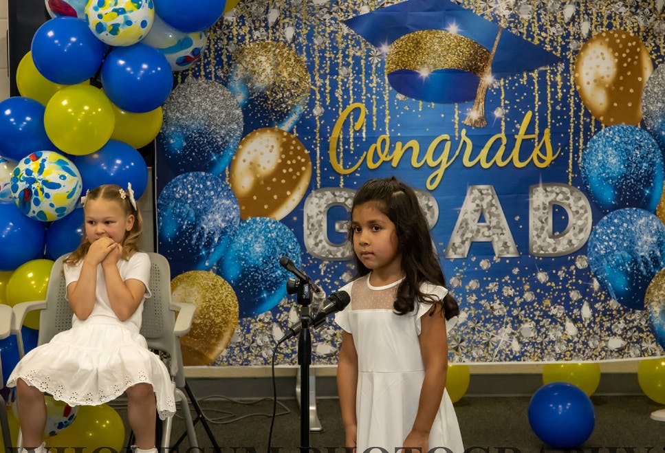 Two children in white dresses, one seated and one standing at a microphone