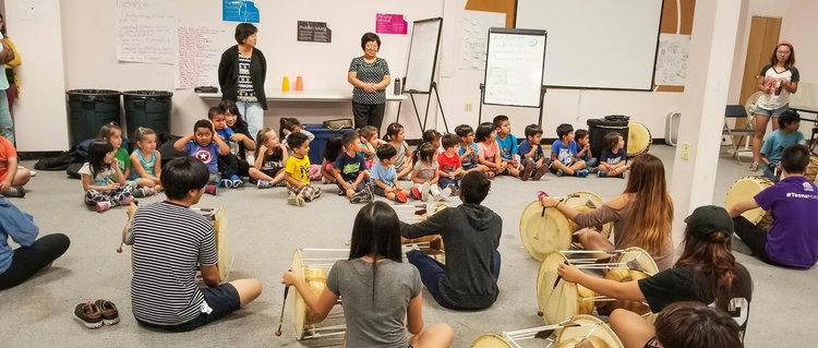 Adults and children sitting on the floor in a circle, some playing drums
