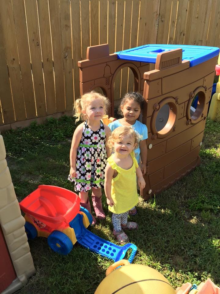 Three children standing in a yard with a playhouse and toy wagon