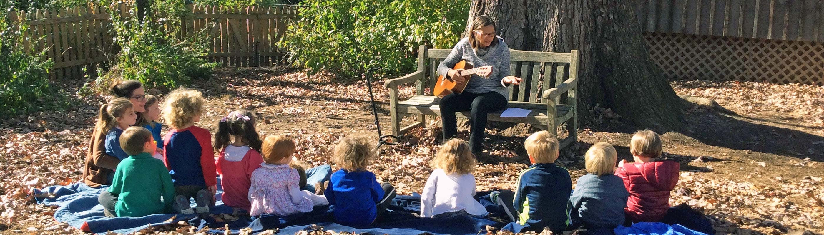Person playing guitar for a group of children sitting on a blanket outdoors