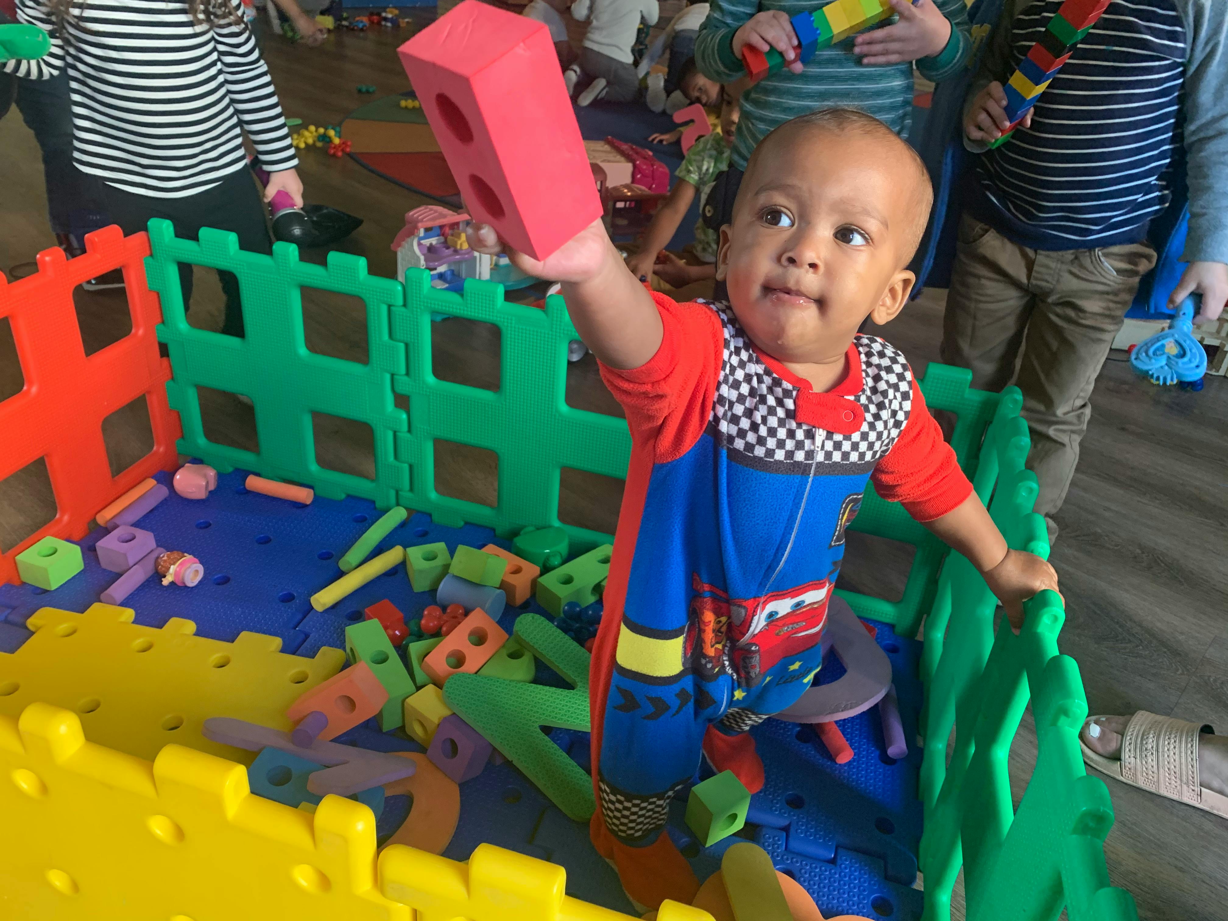 Child holding a red block in a playpen with other blocks