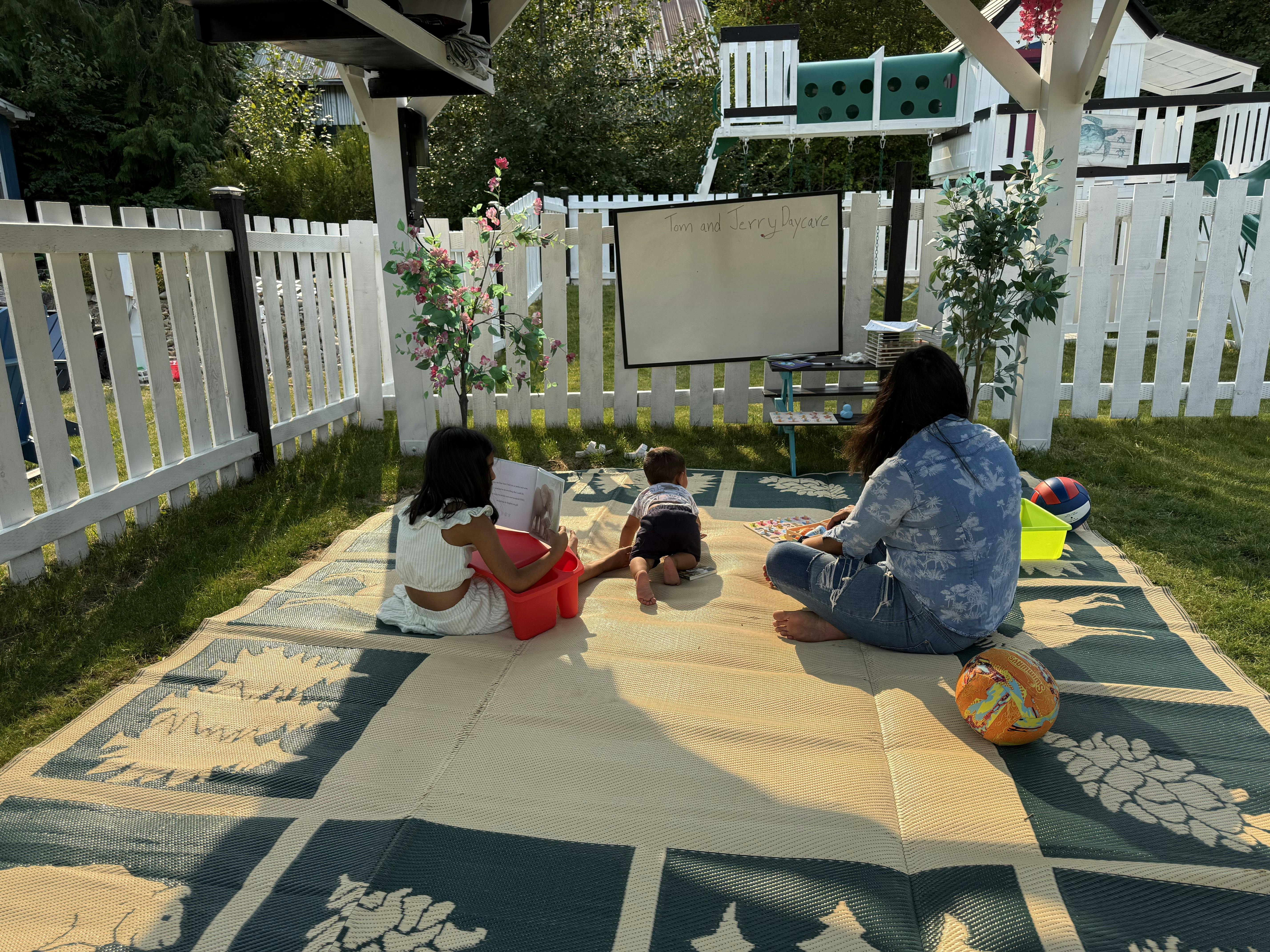 People sitting on a mat in a yard with a whiteboard reading Tom and Jerry Daycare