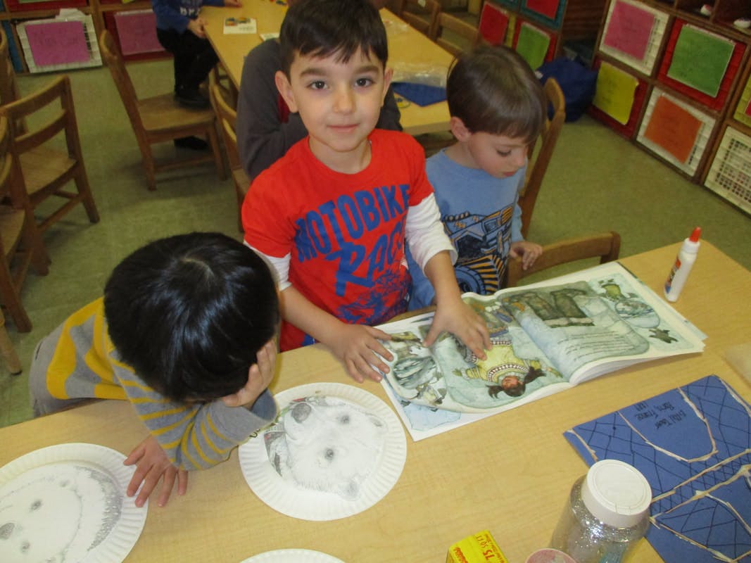 Three children at a table, one looking at a book, another at a paper plate