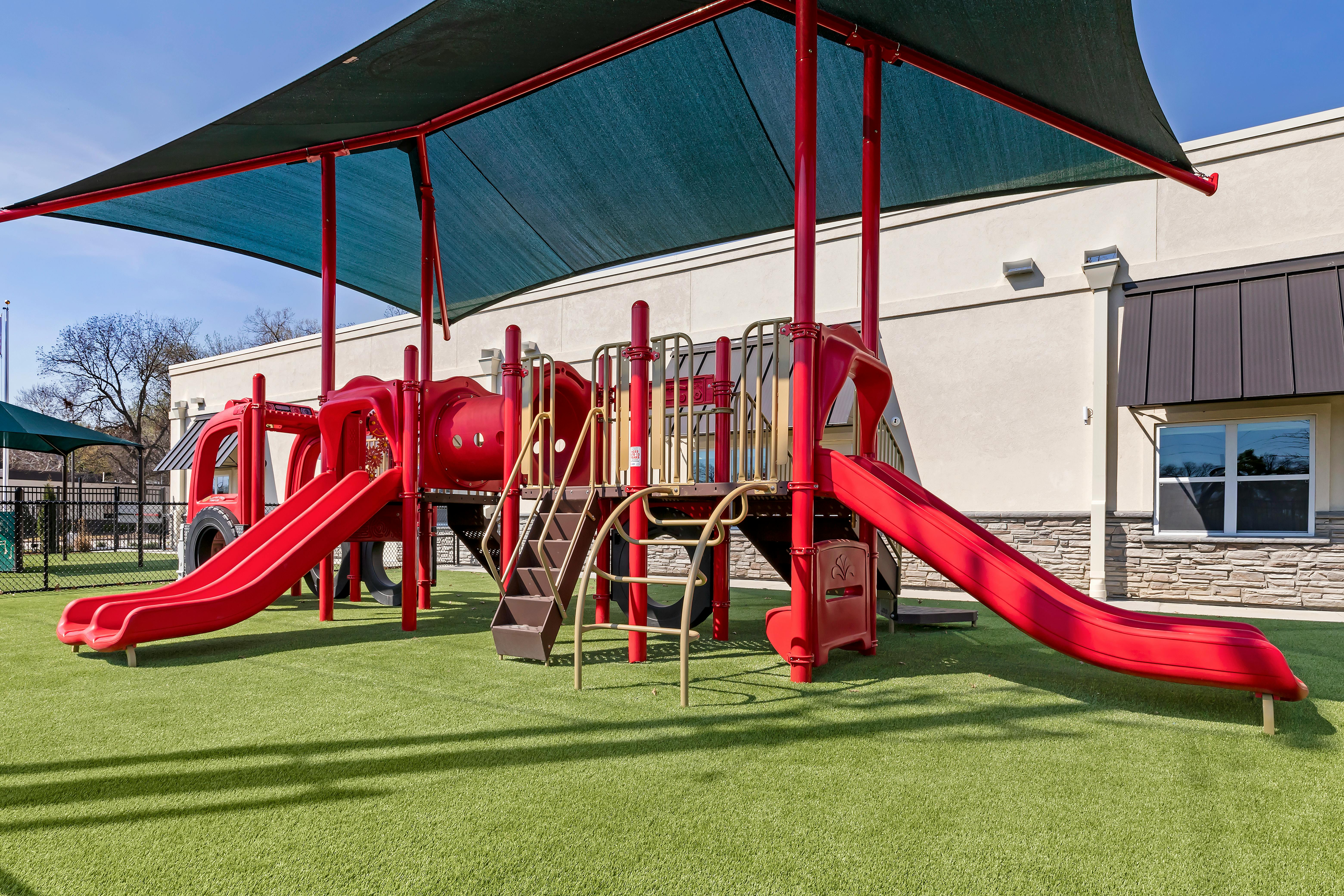 Red playground with slides and stairs on artificial grass, partially shaded