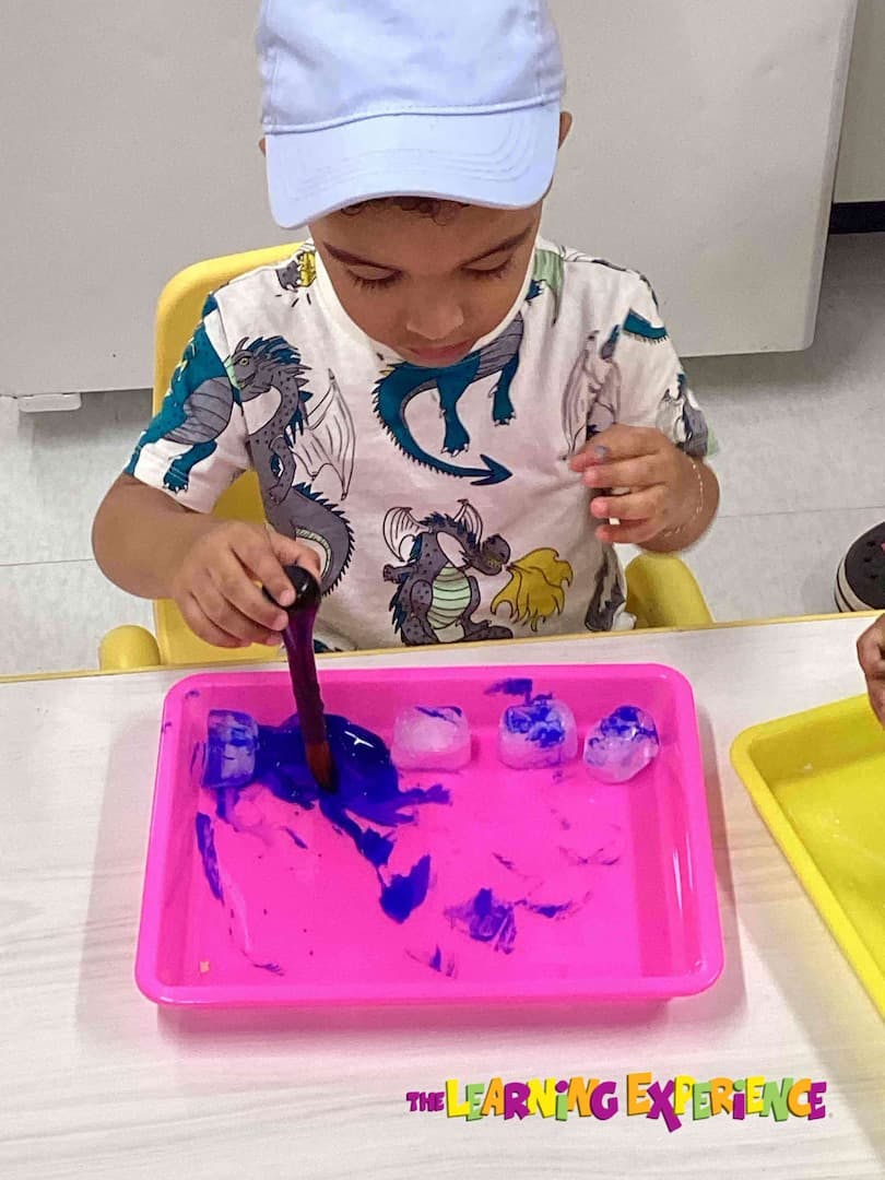 Child in a classroom playing with blue paint and ice cubes in a pink tray