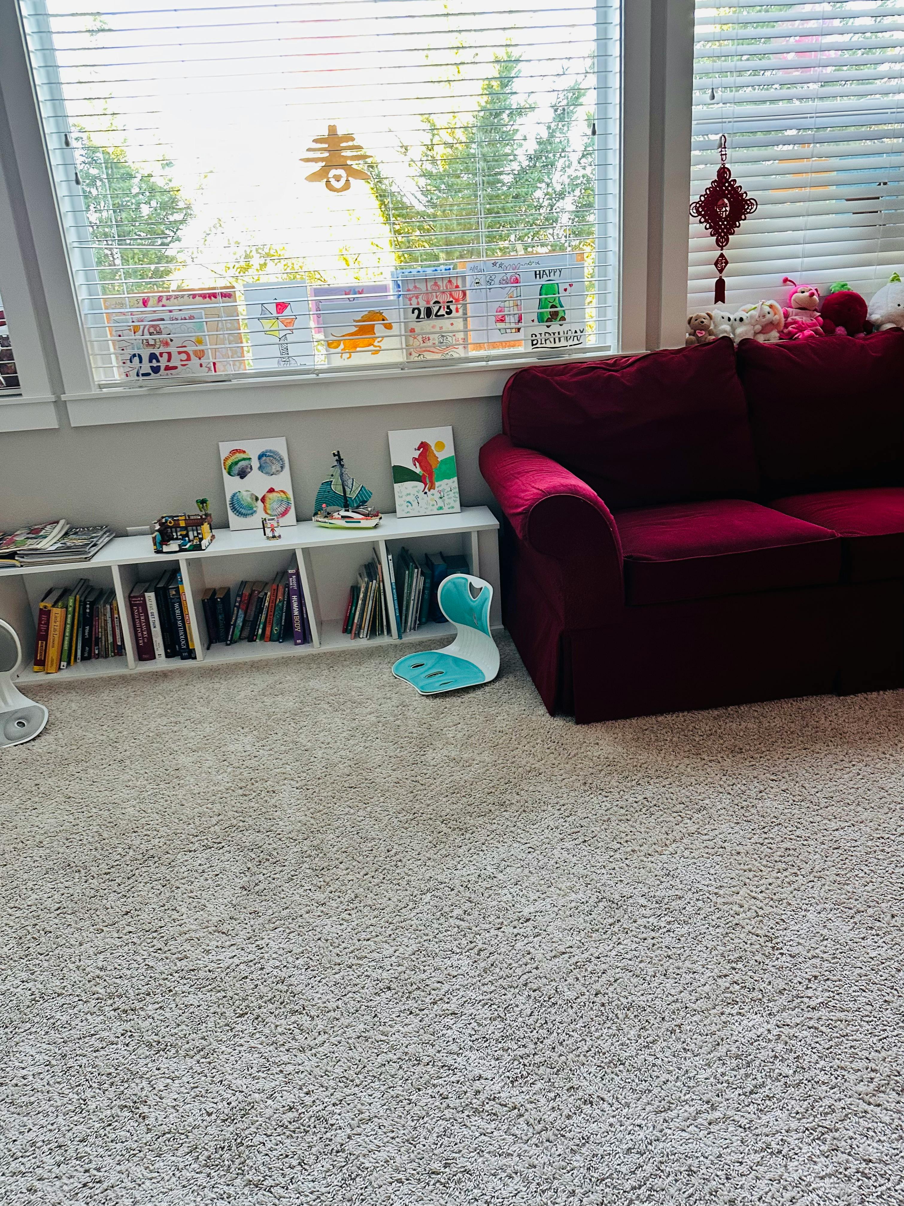 Bookshelf with toys and drawings in front of a window with a red couch