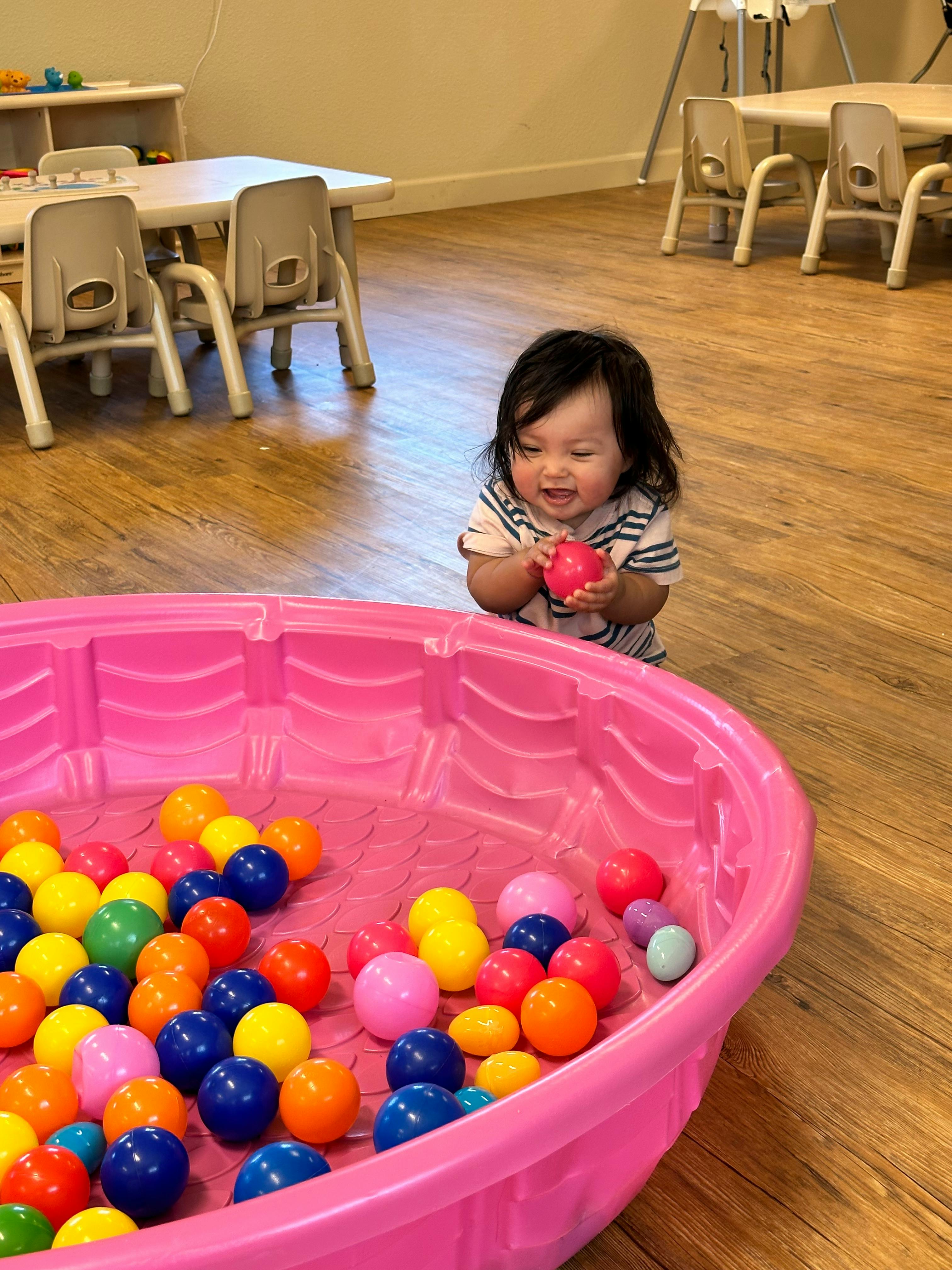 Child holding a ball next to a pink ball pit filled with colorful balls