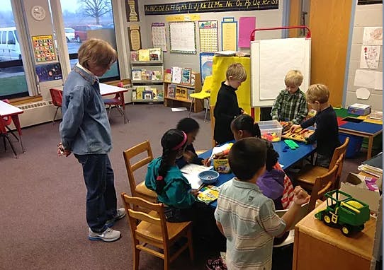 Adult standing near children seated at tables in a classroom