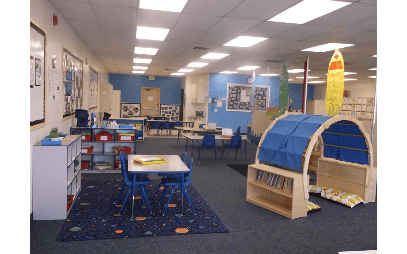 An empty classroom with small tables and chairs, shelves, and a reading nook