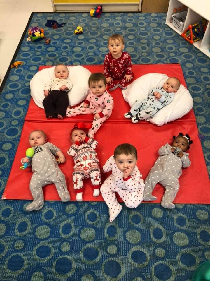 Seven babies on a red mat on a blue patterned carpet