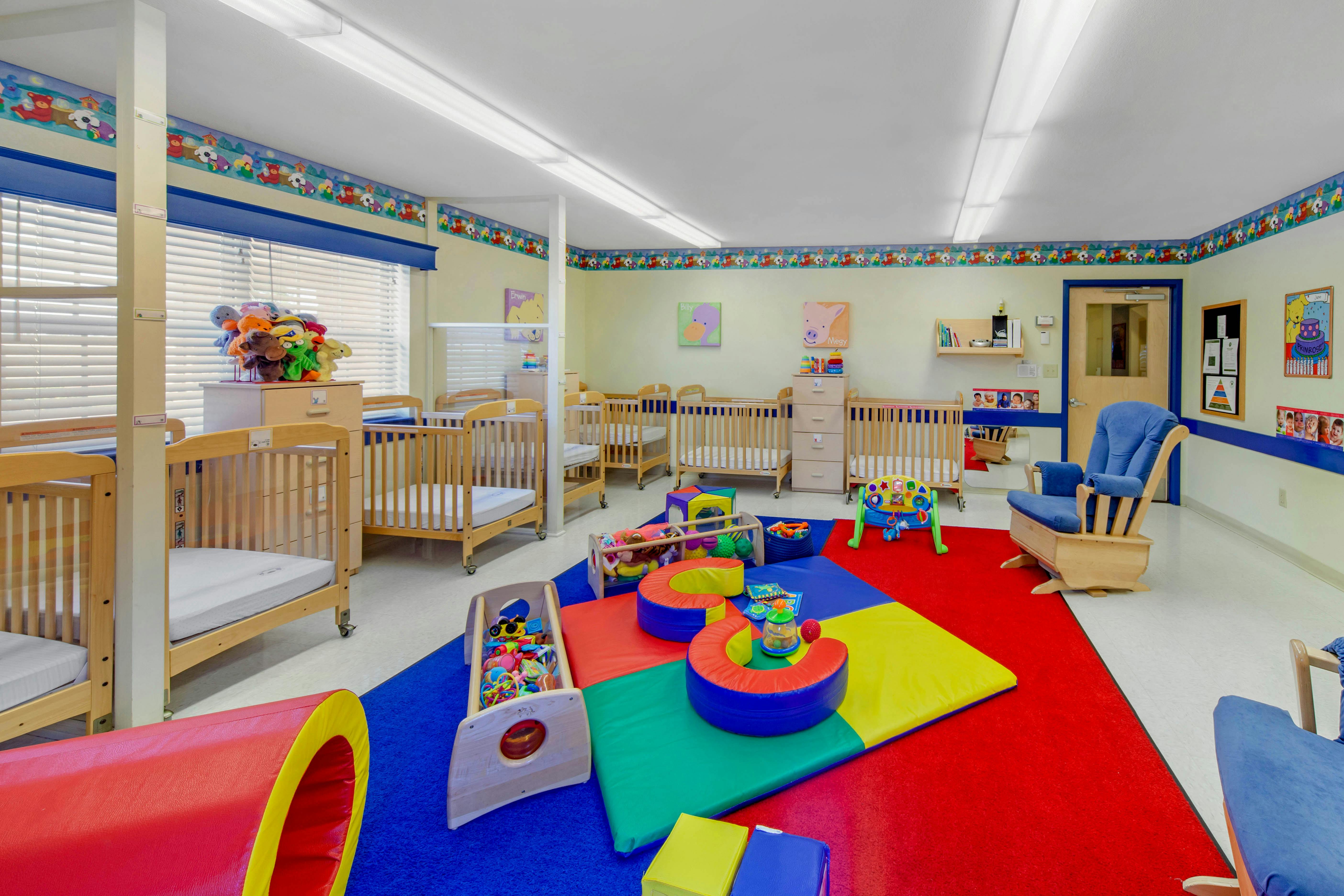 A colorful childcare room with multiple cribs, a red and blue playmat, and toys