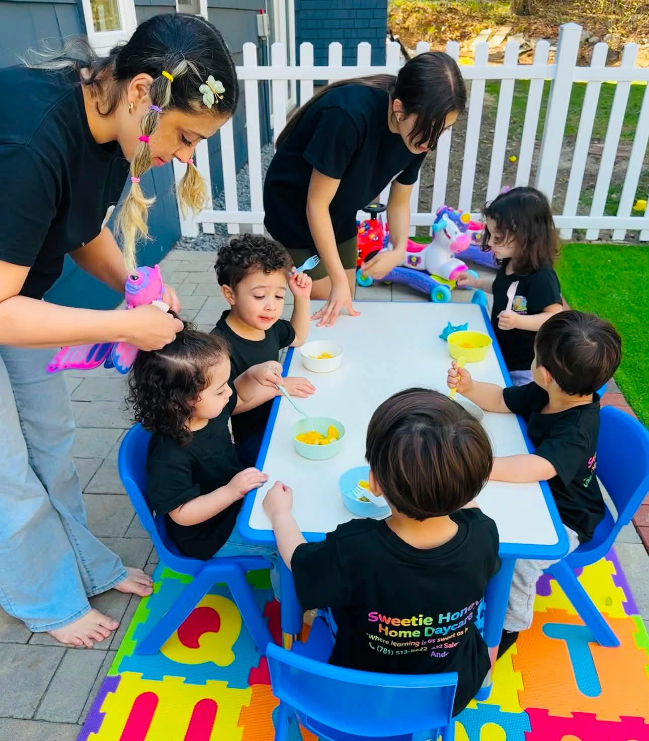 Two people assisting multiple children seated at a small table outdoors, eating snacks