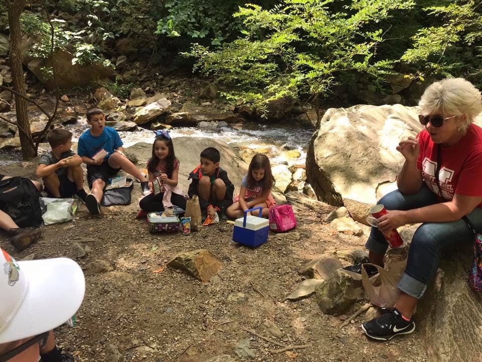 Adult and children sitting on ground near stream with lunchboxes