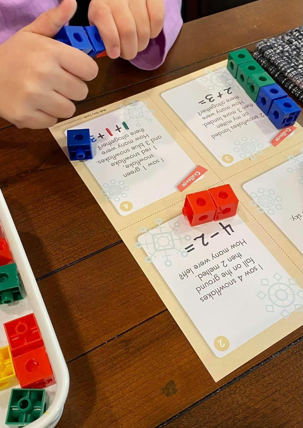 Child's hands holding blue connecting cubes over math problem cards