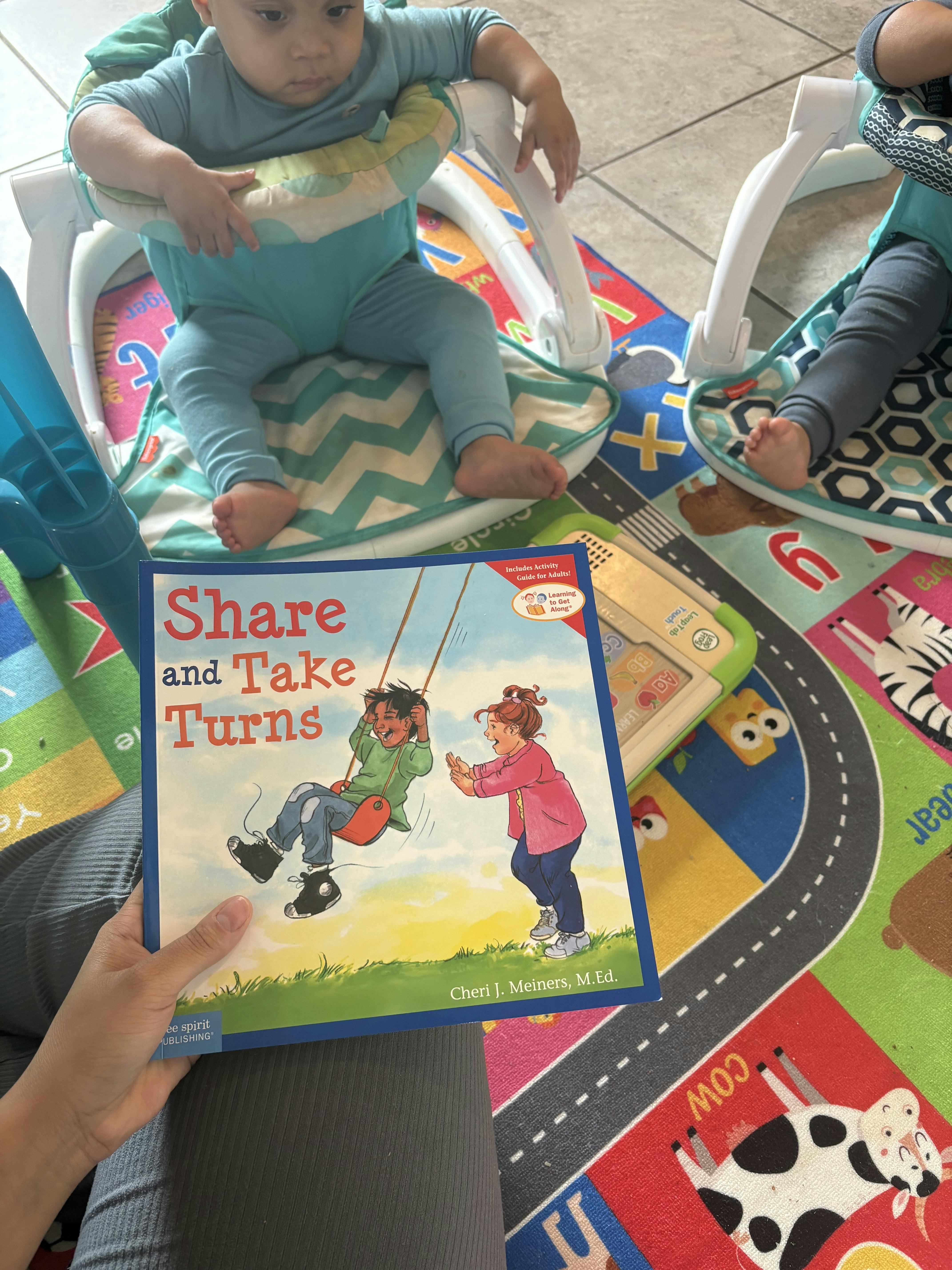 A person holding a book titled Share and Take Turns, two babies in bouncers