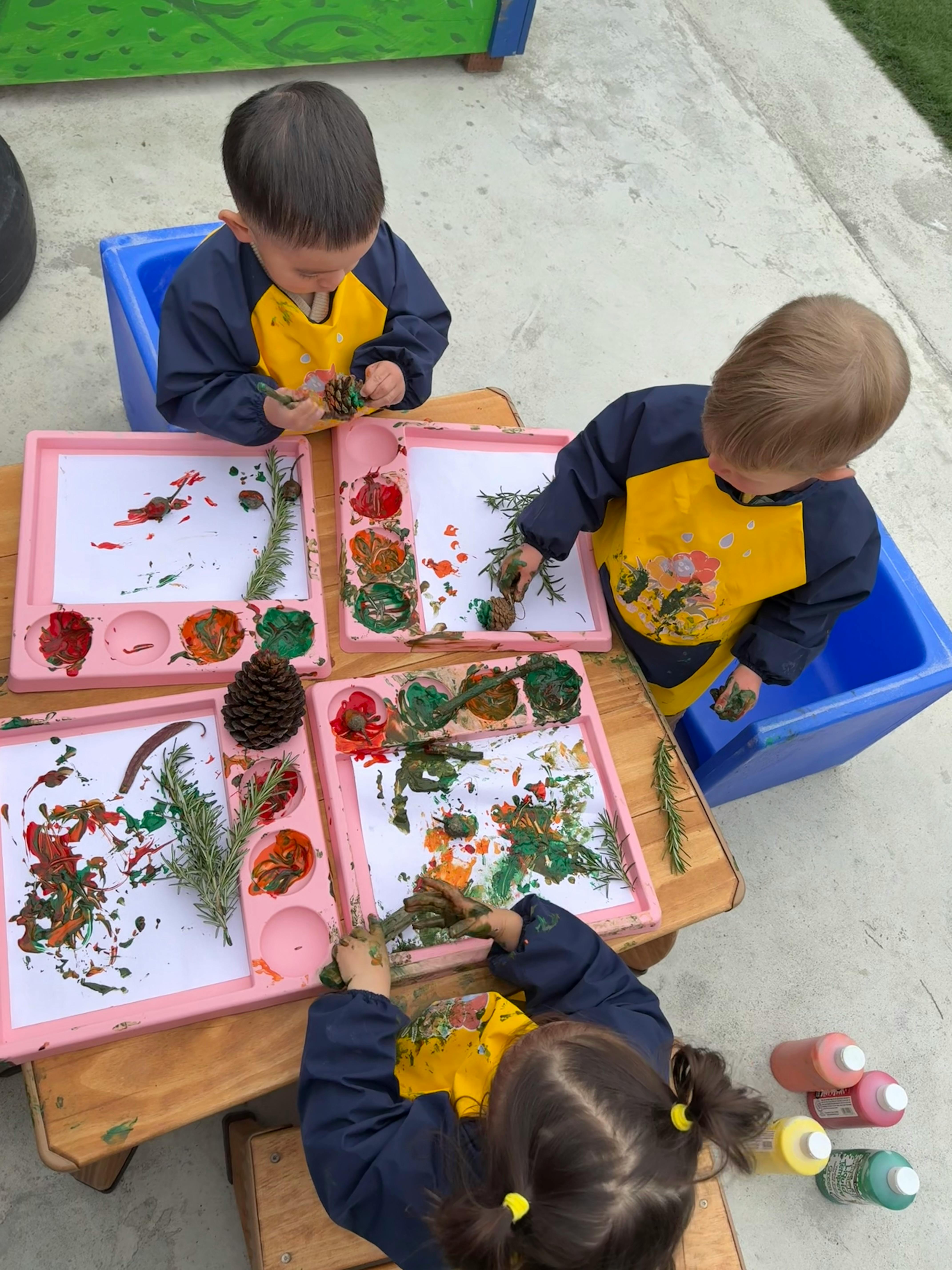 Three children painting with natural elements like pinecones and leaves